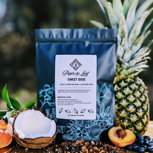 A bag of Piper & Leaf Sweet Dixie Bulk Sachets - 70ct Tea Bags surrounded by a pineapple, coconut, peaches, and berries on a natural backdrop.