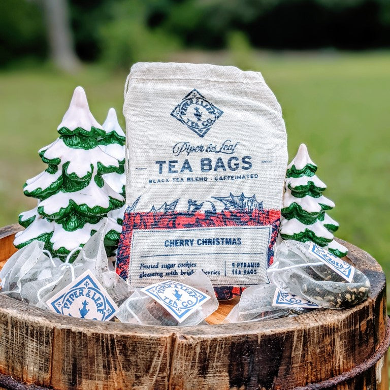 A bag of "Cherry Christmas 9ct Tea Bags in Muslin" from Piper & Leaf Tea Co., surrounded by pyramid tea bags and two small decorative Christmas trees, rests elegantly on a wooden barrel.