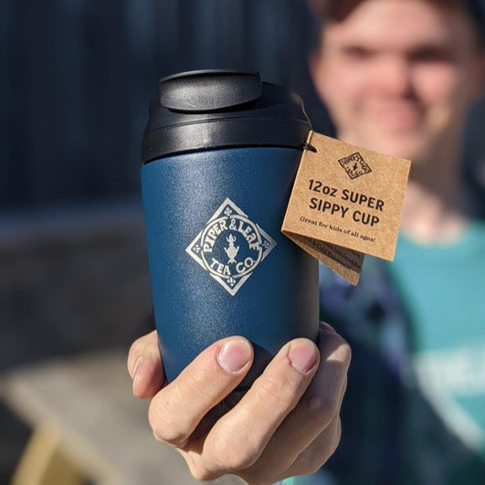 Man in blue hat and plaid shirt holding a blue Piper & Leaf travel cup in right hand, outside in front of building in the sunshine.