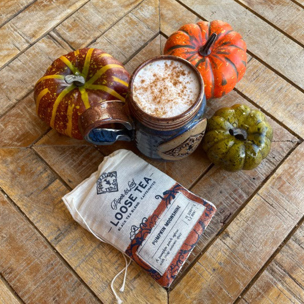 A bag of Pumpkin Moonshine tea and a mug filled with a cinnamon-sprinkled drink surrounded by small pumpkins