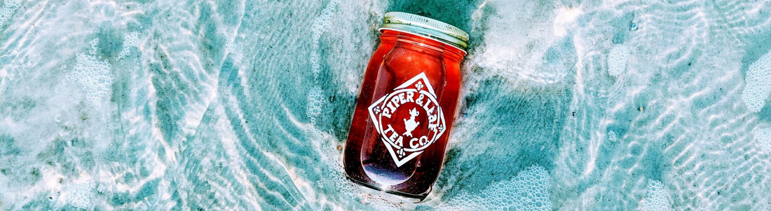 A jar of iced Sweet Dixie, vibrant pink, laying on the ocean's shore amidst the water, glowing with its' vibrant hue against the crystal blue waters