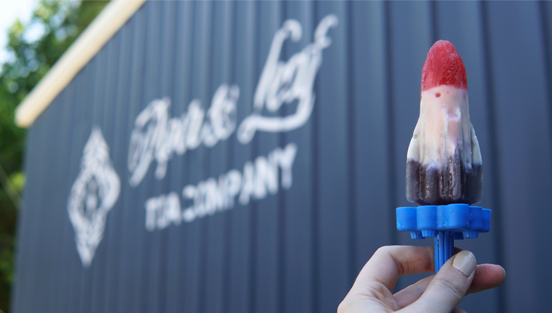 A hand holding a red white and blue rocket pop in front of Pier and Leaf Tea company's warehouse building logo