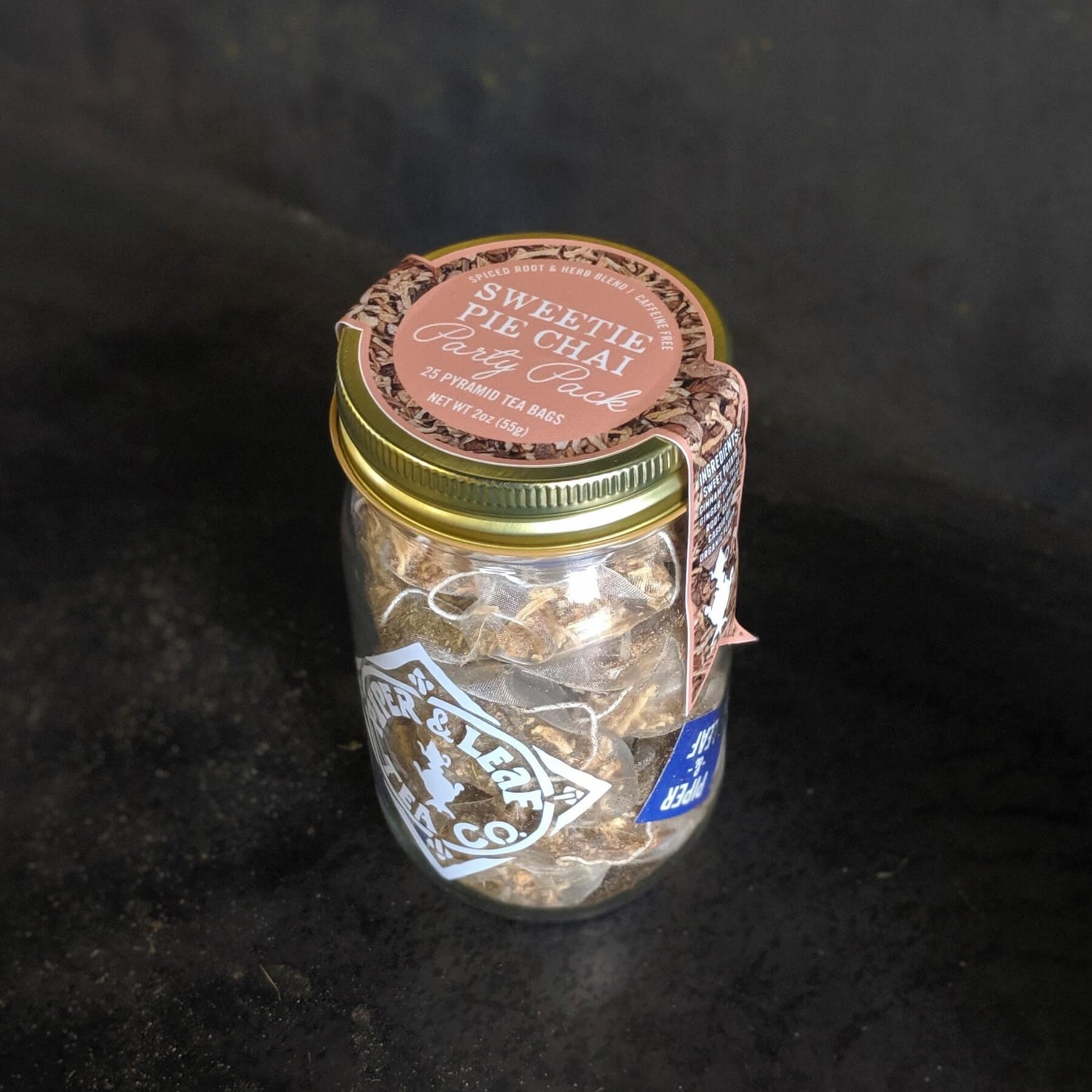 A glass jar with a gold lid labeled "Sweetie Pie Chai Craft Chai" contains chai tea cubes. The jar is placed on a dark surface.