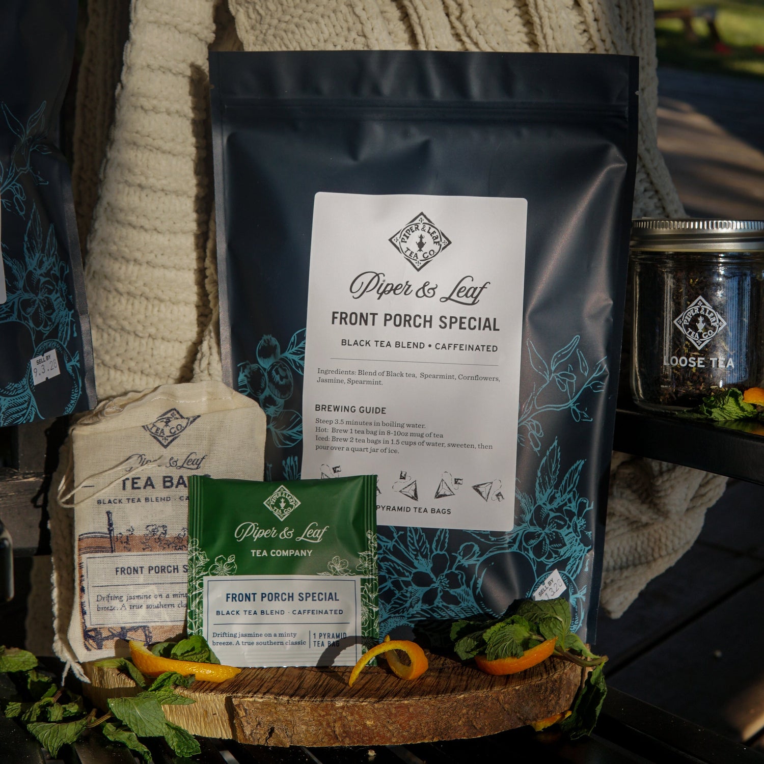 A variety of Piper & Leaf Front Porch Special black tea products, including loose tea, tea bags, and packaging, displayed with fresh mint and orange slices on a wooden surface.