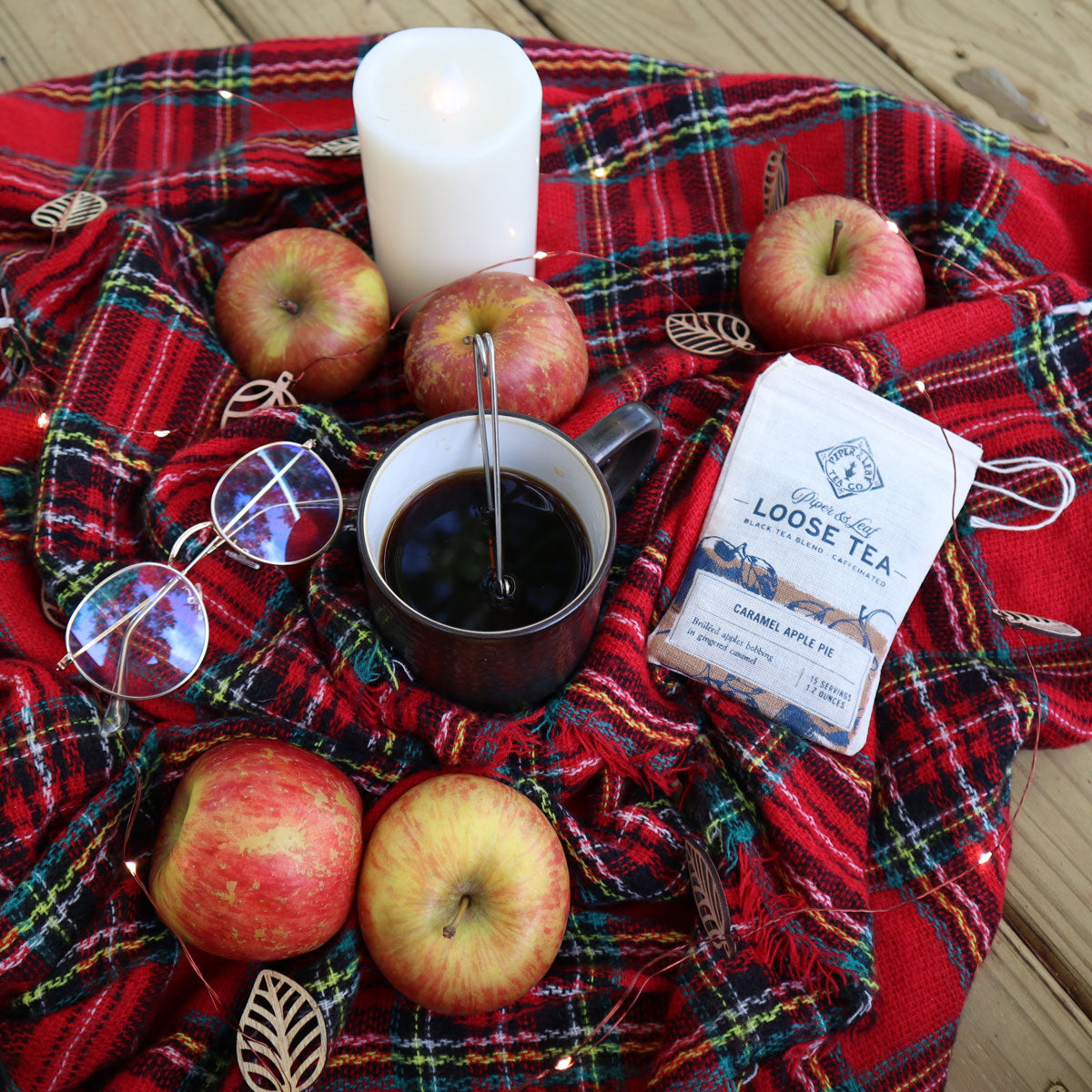 A cozy setup with a cup of tea, apples, glasses, and a candle on a plaid blanket.