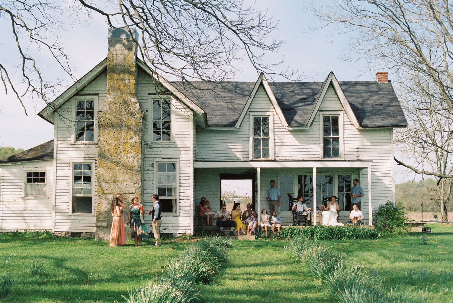 A group of people gather on the porch and lawn of a large, old white house with a stone chimney on a sunny day.