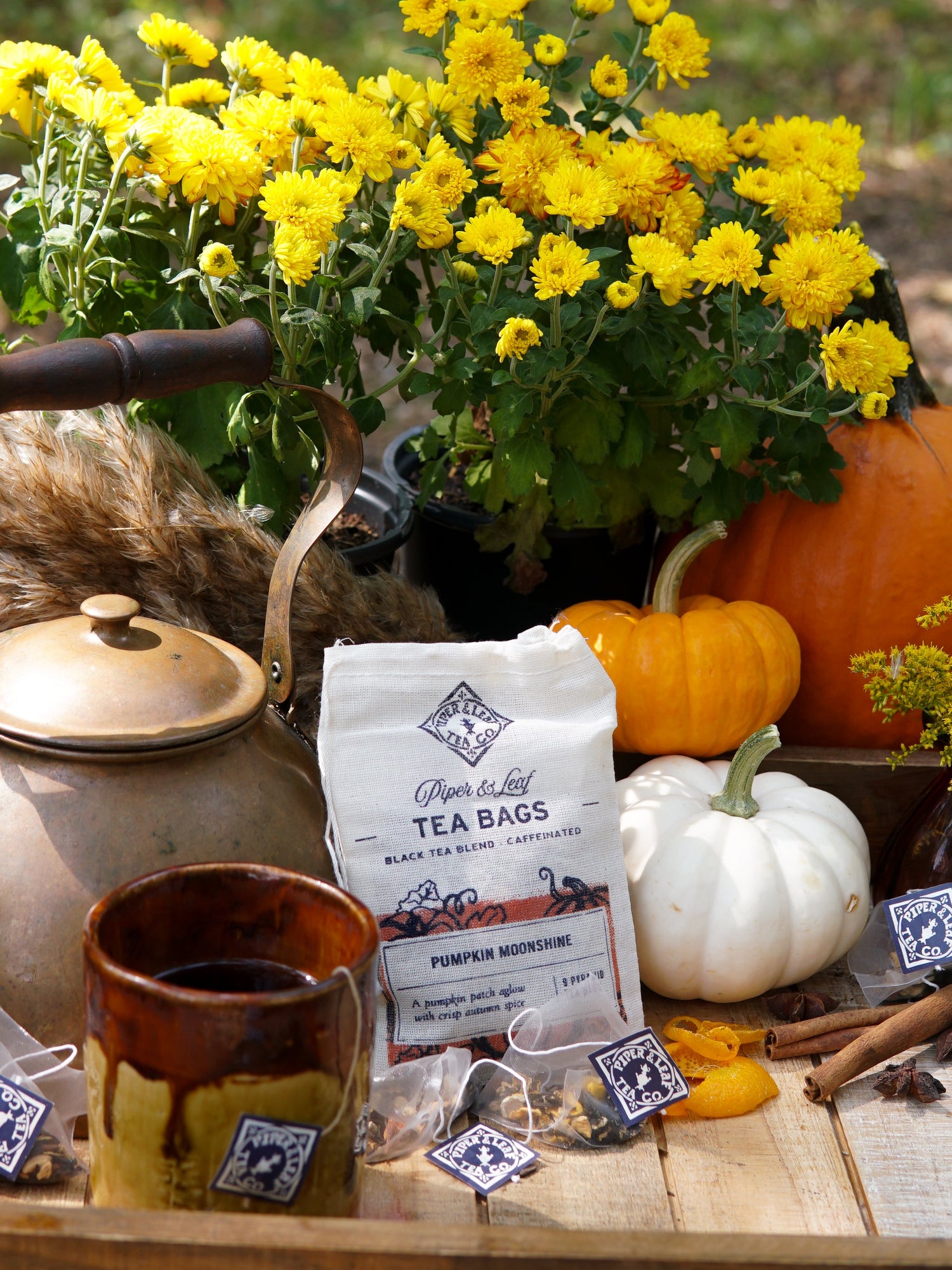 A rustic autumn setup featuring a white mug of tea, a pack of loose tea labeled "Pumpkin Moonshine," two lit white candles, and a pumpkin surrounded by fall leaves on a wooden surface.