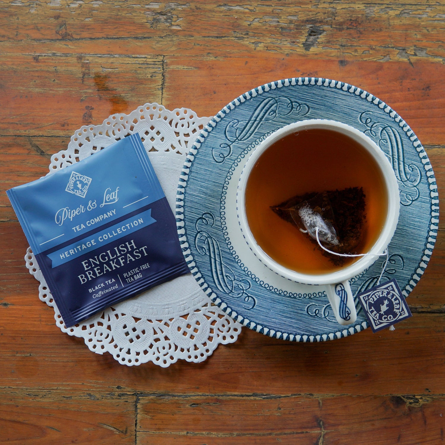 A cup of tea with a tea bag in it sits on a saucer beside a lace doily and an English Breakfast tea packet on a wooden table.