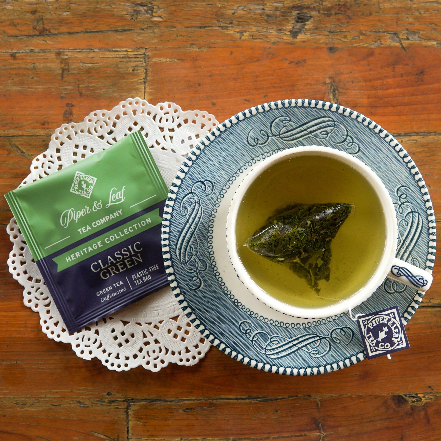 A cup of green tea with a tea bag, a packet labeled “Classic Green” by Piper & Leaf Tea Co., and a paper doily on a wooden table.