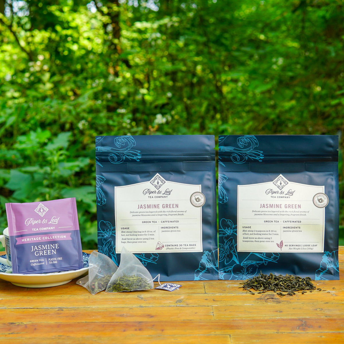 Piper & Leaf Jasmine Green tea products: a box of tea bags, a package of tea bags, and a package of loose-leaf tea, with tea bags and loose tea leaves displayed on a wooden surface.