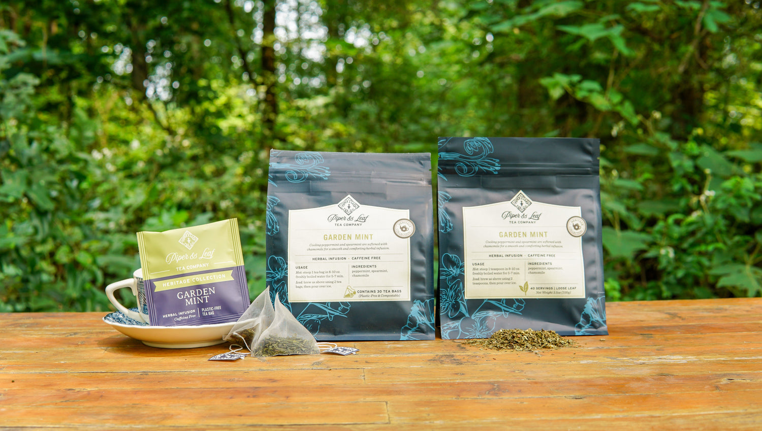 Three packages of Garden Mint tea, one in a cup with pyramid tea bags and dried leaves, are displayed on a wooden table outdoors with green foliage in the background.