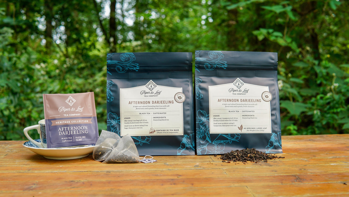 Two bags of Piper & Leaf Afternoon Darjeeling tea, one containing tea bags and one containing loose leaf tea, with a tea bag on a saucer and loose tea scattered on a wooden table.
