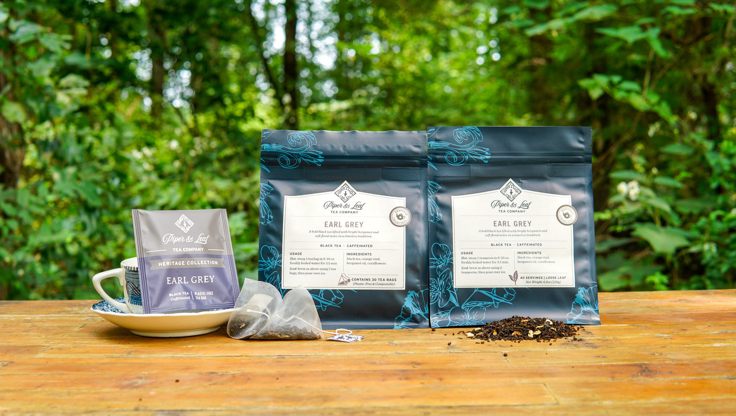 Packets and tea bags of Earl Grey tea are arranged on a wooden table outdoors with loose tea, a teacup, and a green leafy background.