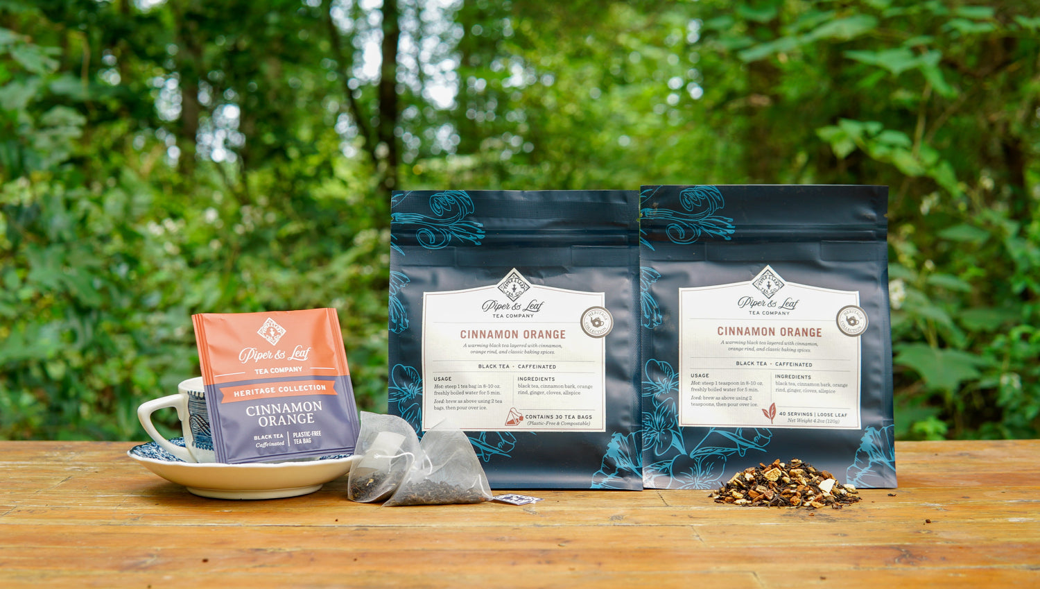 Two bags and one sachet of Cinnamon Orange black tea are on a wooden table outdoors, along with a white cup, saucer, tea bag, and loose tea leaves. Green foliage is visible in the background.