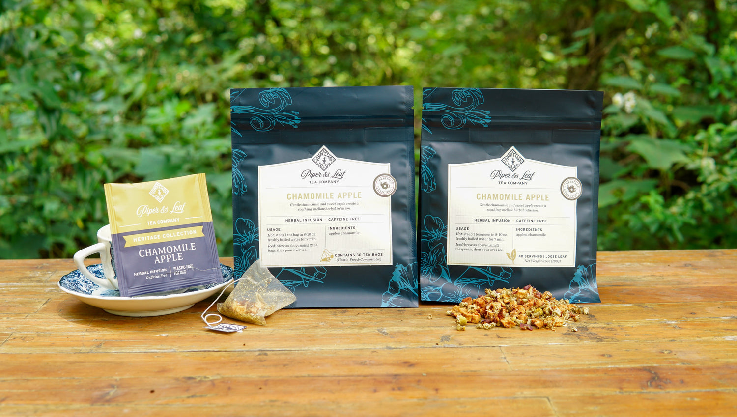 Two bags, a sachet, and a cup of Piper & Leaf Chamomile Apple tea are on a wooden table outdoors, with loose tea leaves and a green leafy background visible.