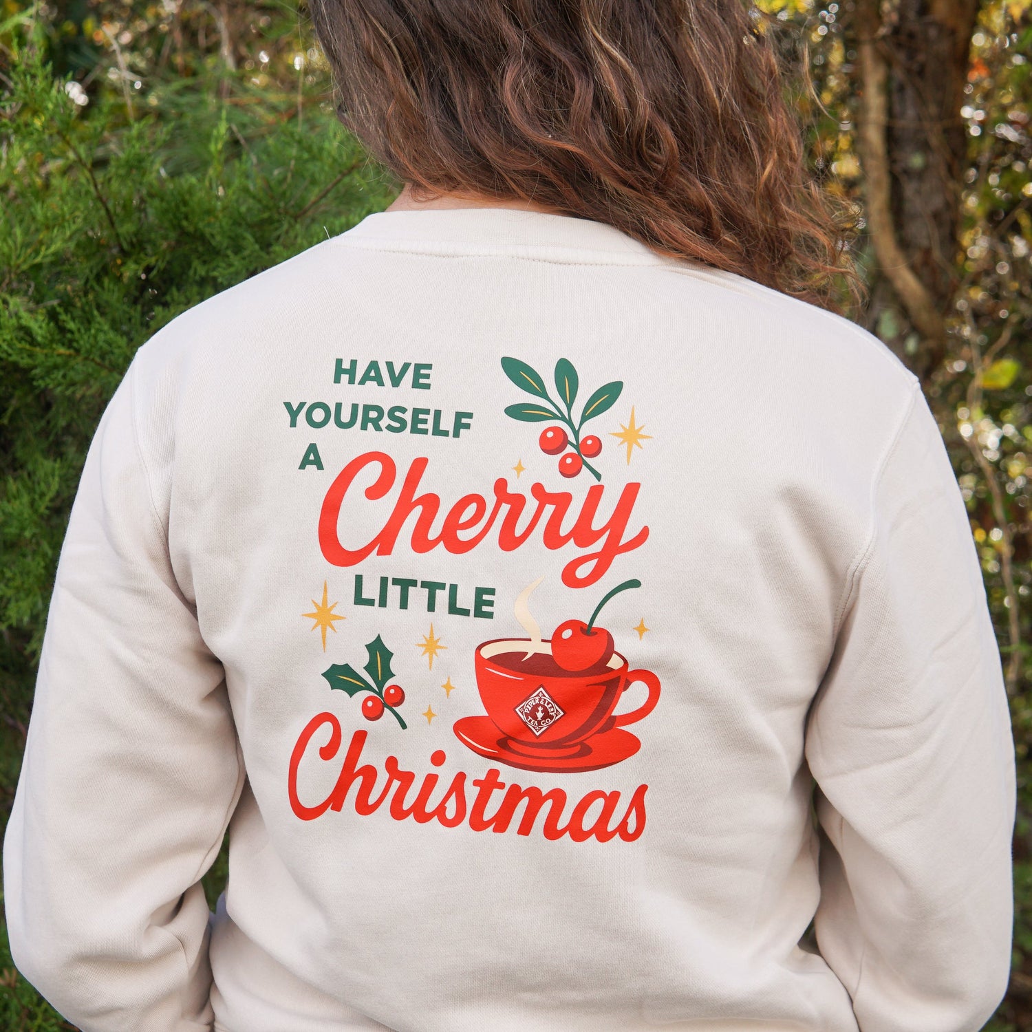 Person wearing a cream sweatshirt with festive text and a cherry teacup design reading, “Have Yourself a Cherry Little Christmas.” Background shows greenery and trees.