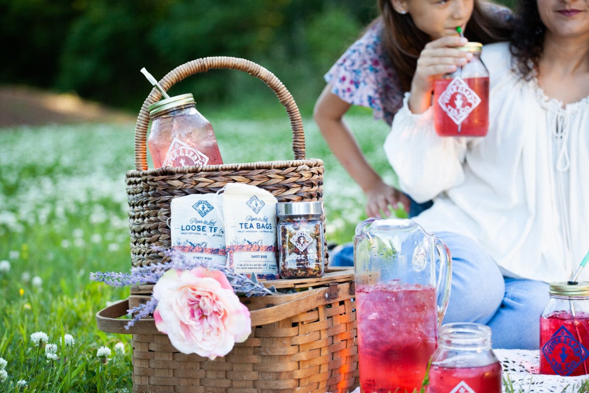 A picnic scene with jars of iced tea, a woven basket, tea containers, a flower, and two people sitting on the grass, one drinking from a mason jar with a straw.