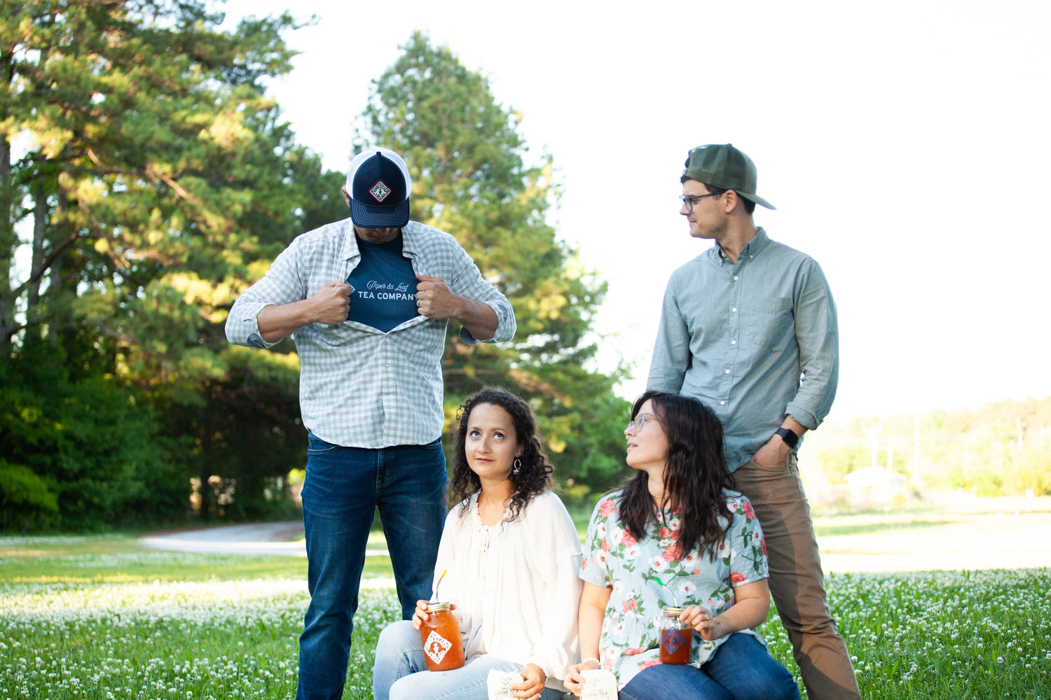 A group of four people is outdoors, three sitting with drinks and one standing, showing off a T-shirt. Trees and greenery are in the background.