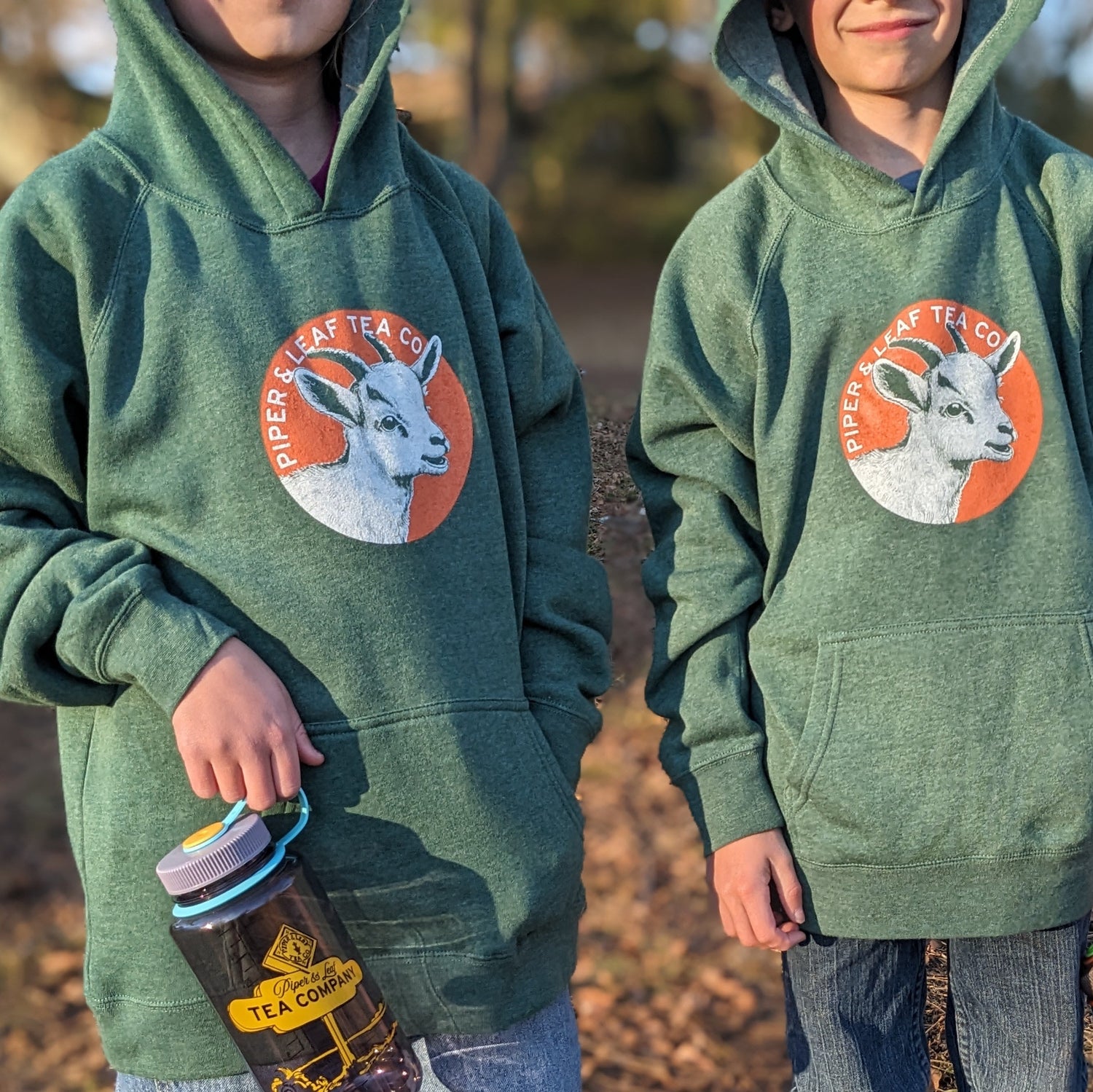 Two individuals in matching green hoodies with a goat logo, one holding a jar of tea.