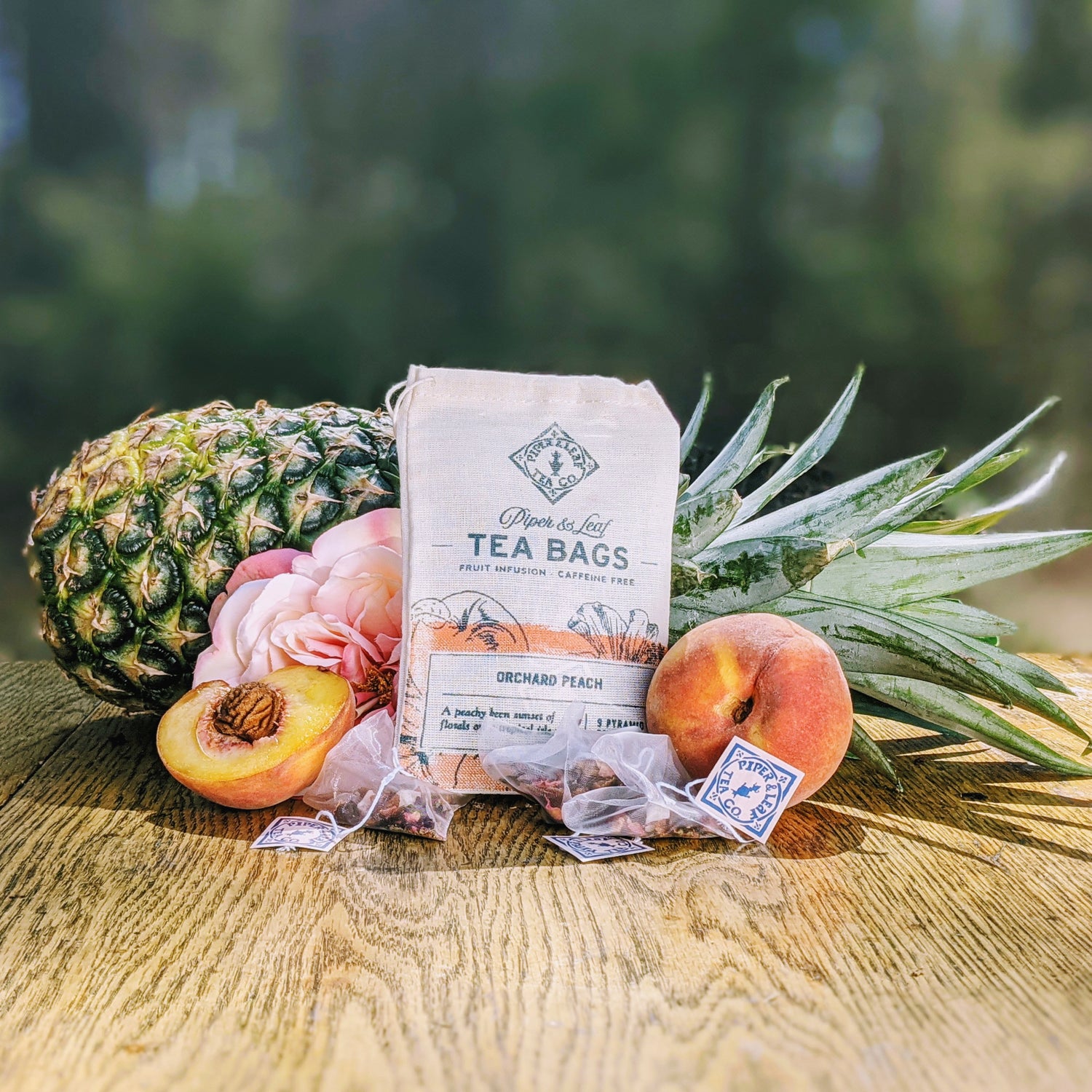 A package of orchard peach tea bags is displayed on a wooden surface with a pineapple, peaches, and a pink flower in the background. Tea bags and loose fruit are arranged in front.