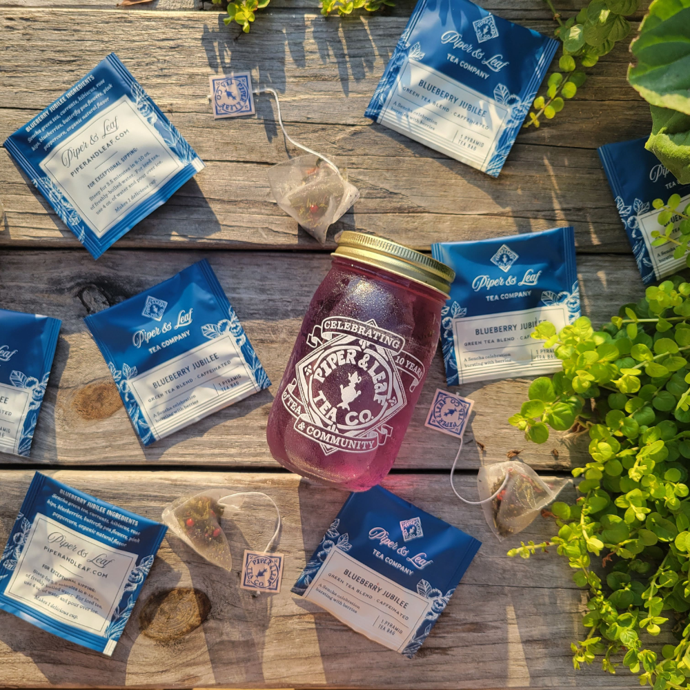 A mason jar with purple liquid is surrounded by scattered blueberry herbal tea packets and tea bags on a rustic wooden surface. Bright green plants frame the scene.