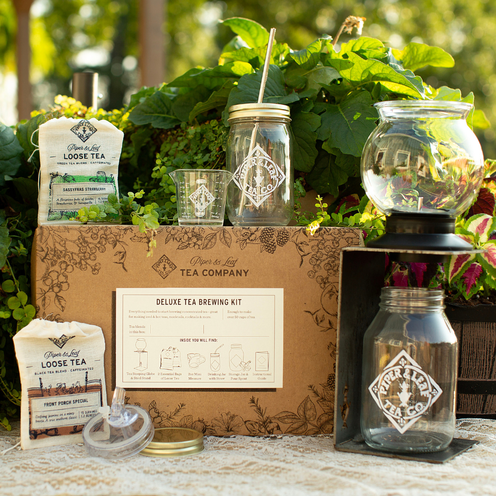 A deluxe tea brewing kit with tea bags, a glass jar with straw, a glass infuser, and a branded box displayed outdoors with green plants in the background.