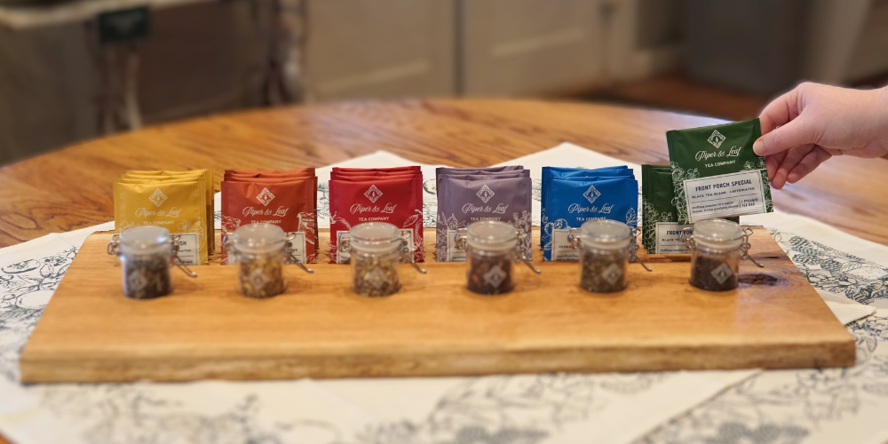 Assorted tea packets in various colors arranged on a wooden board with small jars in front. A hand holds a green packet labeled "First Touch Special.