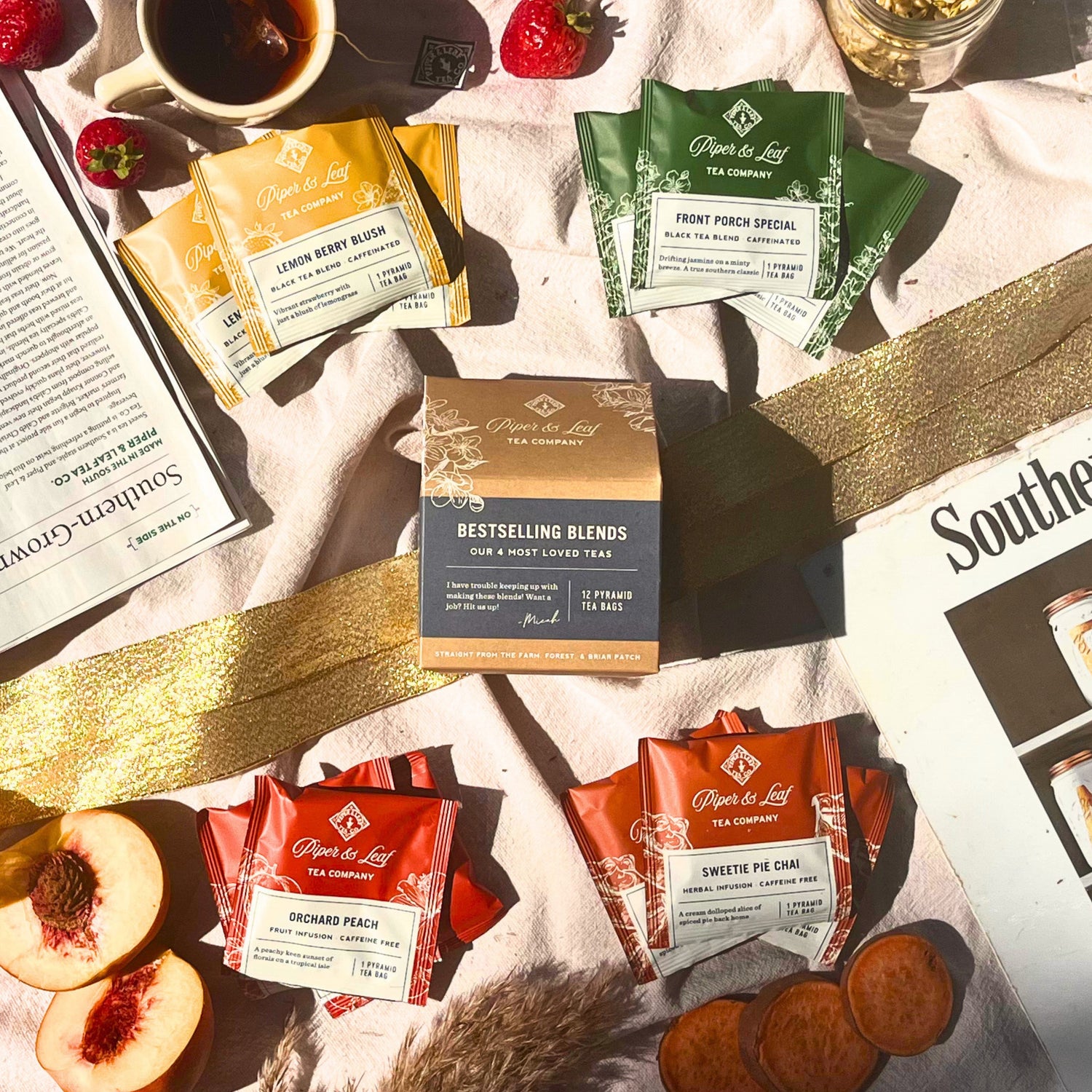 Assorted Piper & Leaf tea packets are arranged on a picnic blanket with sliced peaches, strawberries, a drink, and newspapers in natural sunlight.