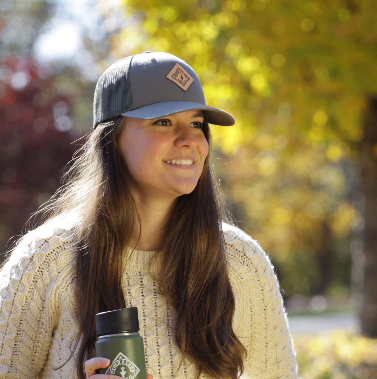 A woman in a knit sweater and baseball cap stands outdoors, holding a reusable bottle, with autumn trees in the background.