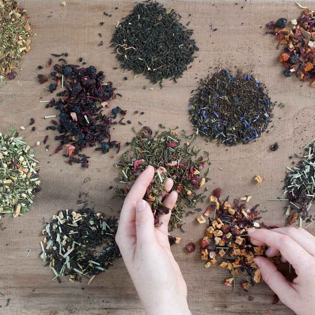 Various loose-leaf teas and botanicals arranged on a surface with a person's hands interacting with them.