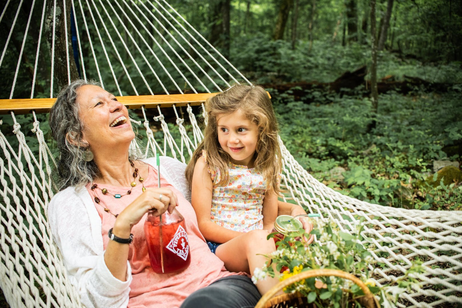 An elderly woman and a young girl laughing together in a hammock in a forest, with a basket and drinks nearby.