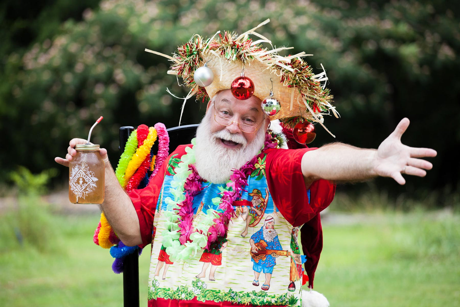 A person dressed in festive attire, including a hat decorated with tinsel and ornaments, a colorful lei, and holding a drink, extends one arm outward while standing outdoors.