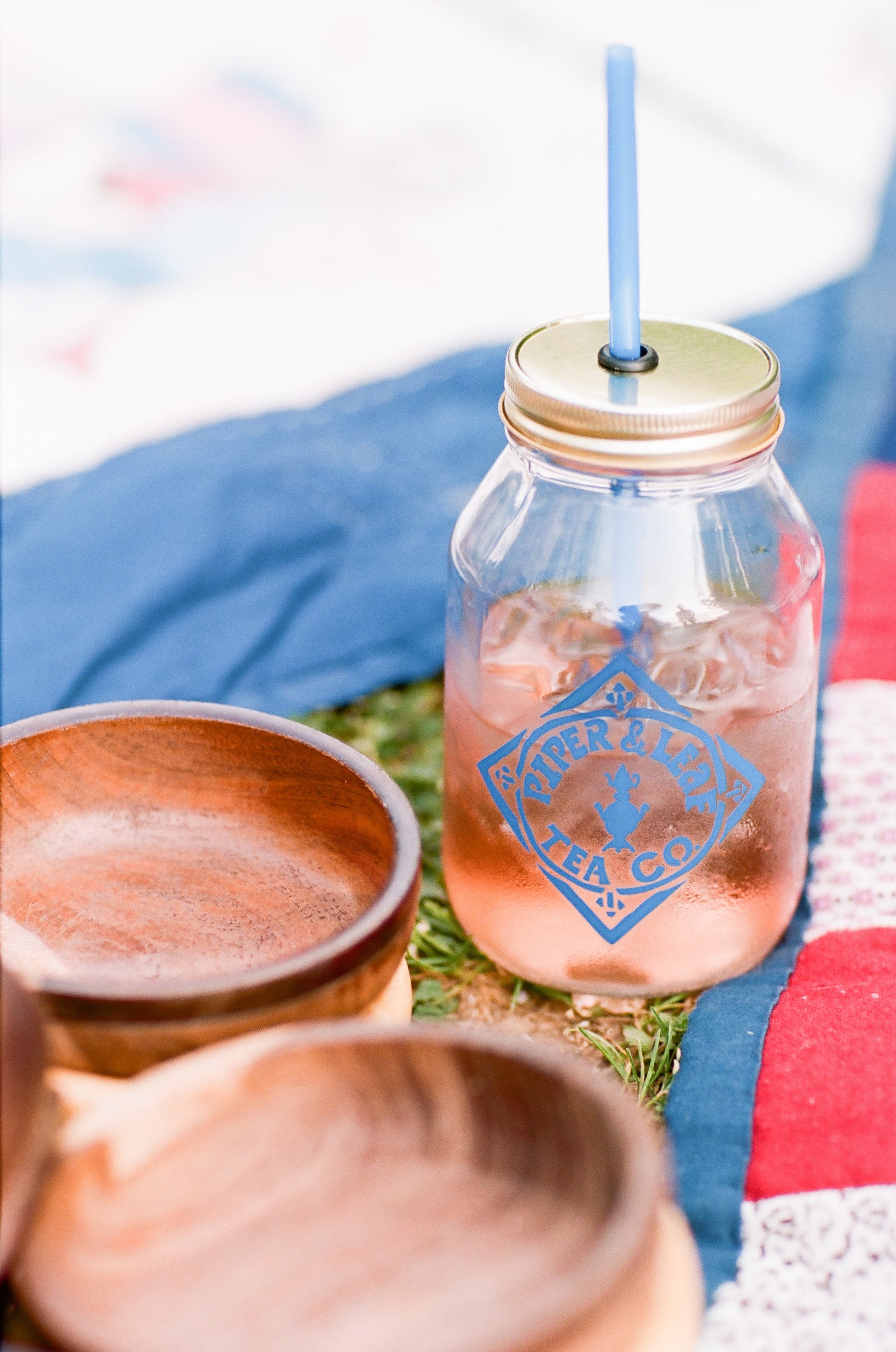 A Signature Mason Drinking Jar - Quart Size with a straw sits on a picnic blanket, ready to be filled with refreshing iced tea from Piper & Leaf Tea Co.