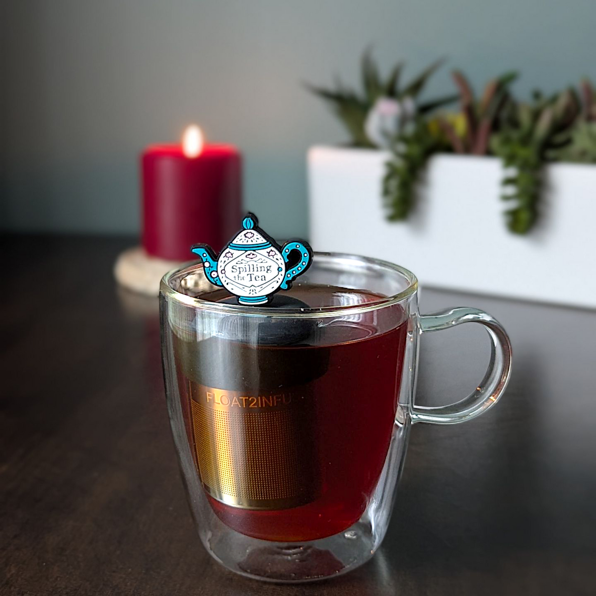 A glass mug of tea featuring the Float2infuse Tea Infuser’s Spill the Tea - Floating Tea Infuser, with its stainless steel mesh design and whimsical teapot-shaped tag, sits on a dark table beside a glowing red candle and a white succulent planter—an inviting scene to enjoy your favorite tea accessory.
