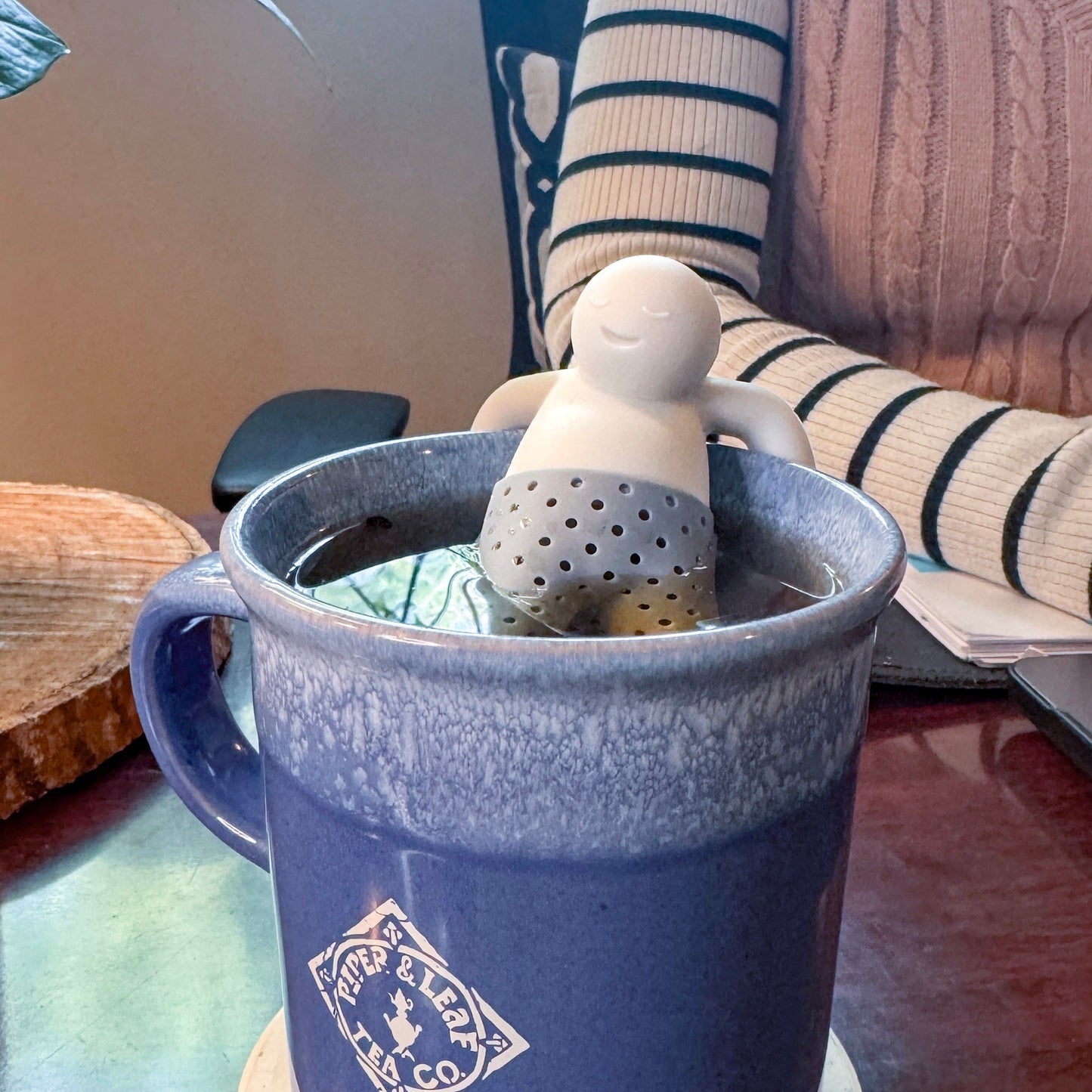 A mug of loose-leaf tea is brewed with the Piper & Leaf Tea Co. Mr. Tea Infuser, a BPA-free device shaped like a relaxed human figure floating inside; someone in a striped sweater sits in the background.