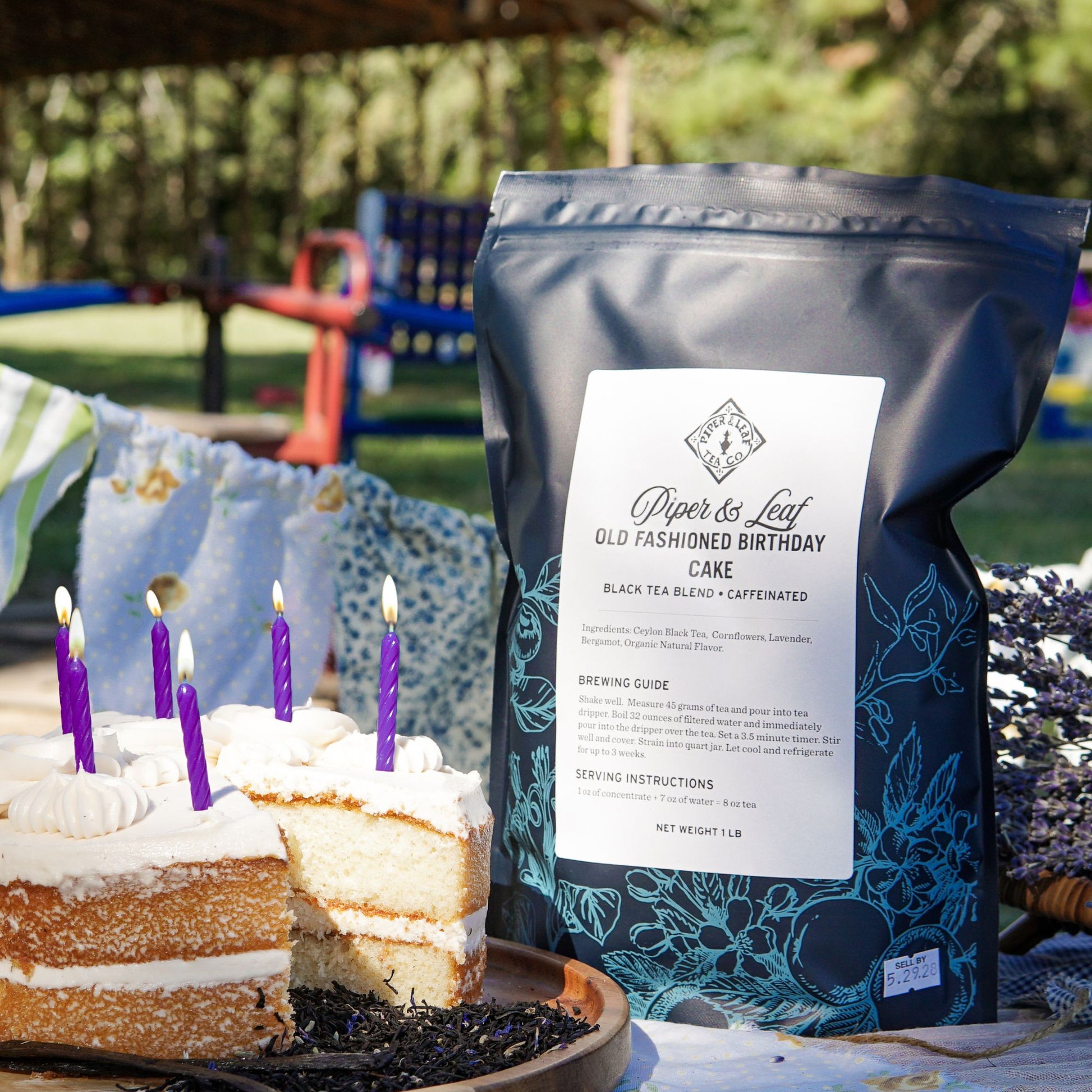 A Piper & Leaf Tea Co. Old Fashioned Birthday Cake Pound Bag (190 servings) sits by a birthday cake with lit candles and a missing slice on an outdoor table.