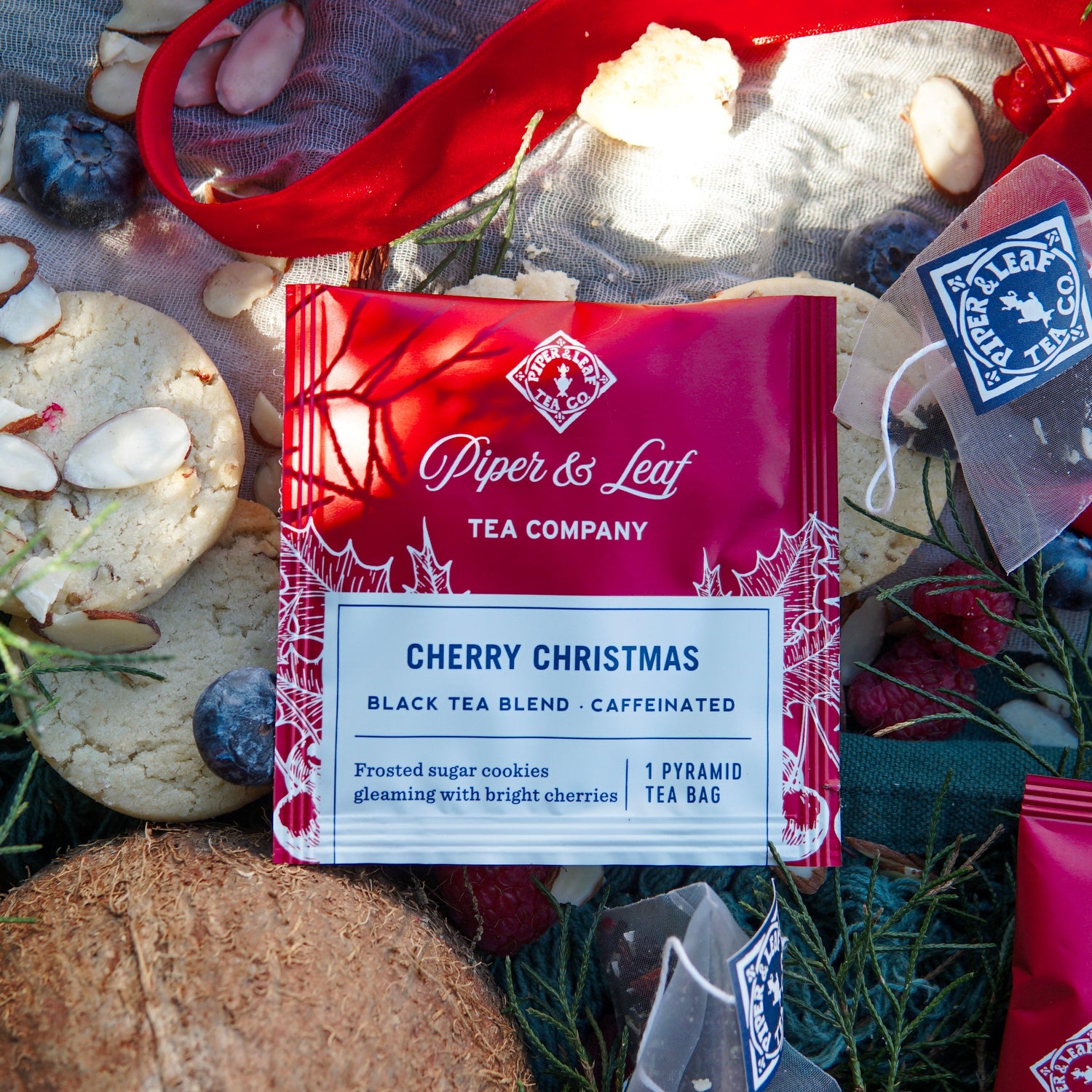 A Piper & Leaf Tea Co. Cherry Christmas Case of Individually Wrapped Tea Bags (50ct) is displayed on a festive background, surrounded by cookies, berries, coconut, and single-serve tea sachets.