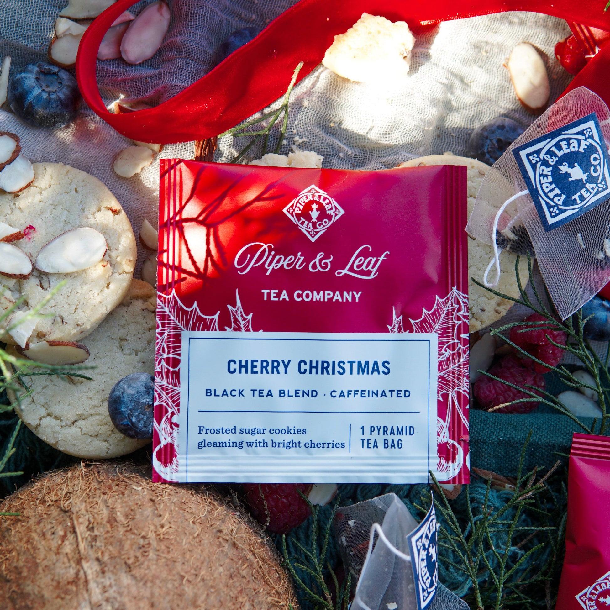 A Piper & Leaf Tea Co. Cherry Christmas Case of Individually Wrapped Tea Bags (50ct) is displayed on a festive background, surrounded by cookies, berries, coconut, and single-serve tea sachets.