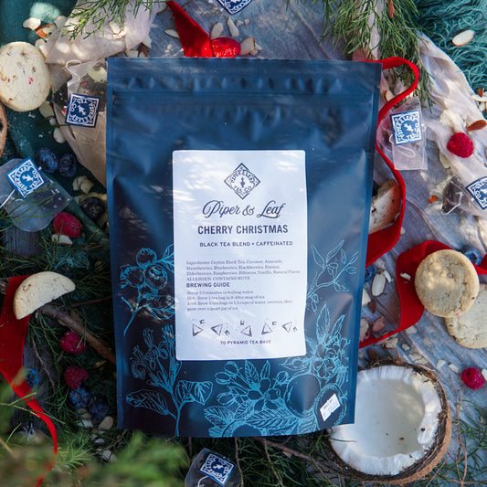 A bag of Piper & Leaf Tea Co. Cherry Christmas Bulk Sachets (70ct tea bags) rests on a festive surface surrounded by cookies, ribbons, greenery, and loose tea pyramids.