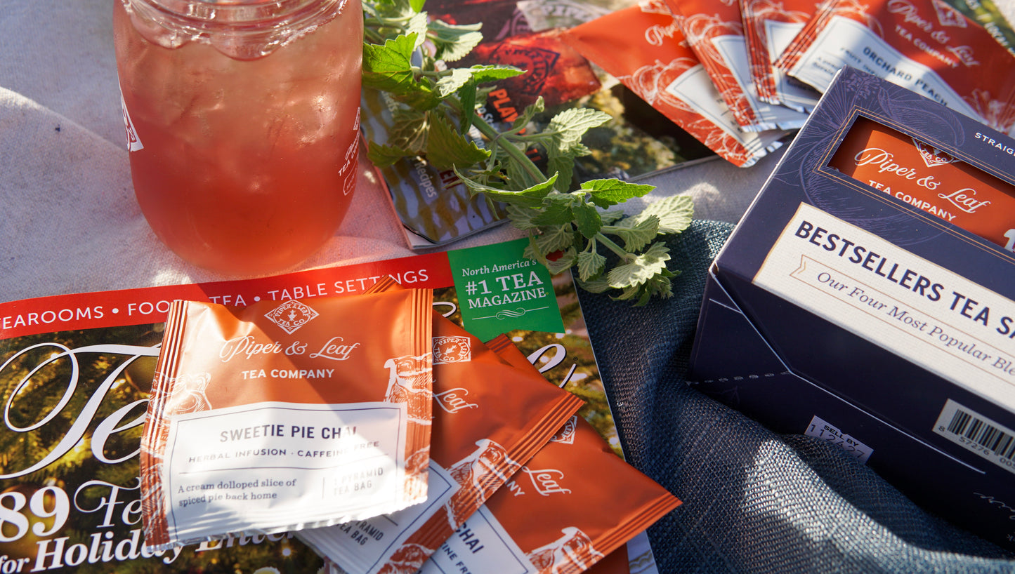 A mason jar of iced tea, a Piper & Leaf Tea Co. Bestselling Tea Sampler (box of 16 tea bags), assorted tea packets, mint sprigs, and a tea magazine are displayed on a sunlit table.