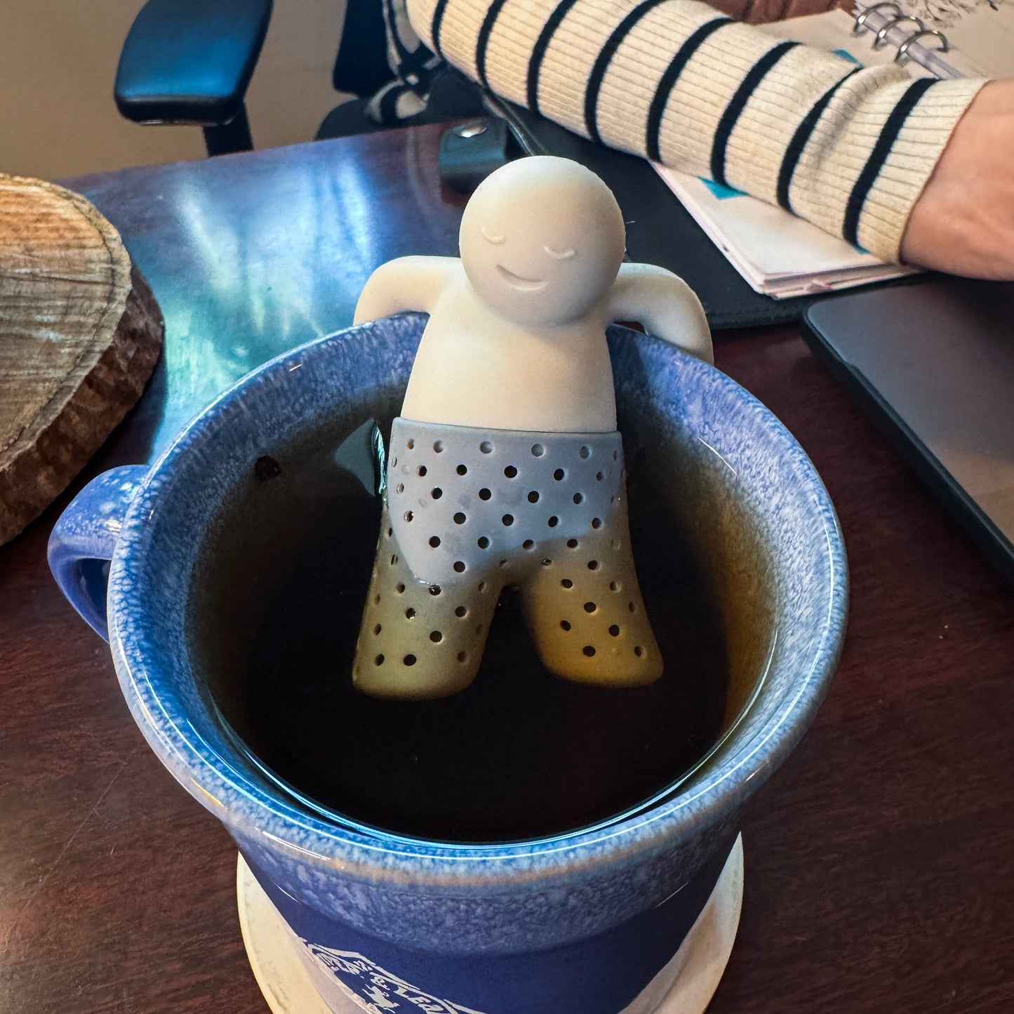 A Piper & Leaf Tea Co. Mr. Tea Infuser, BPA-free and shaped like a relaxed person, steeps loose-leaf tea in a blue mug on a desk beside an arm, notebook, and laptop.