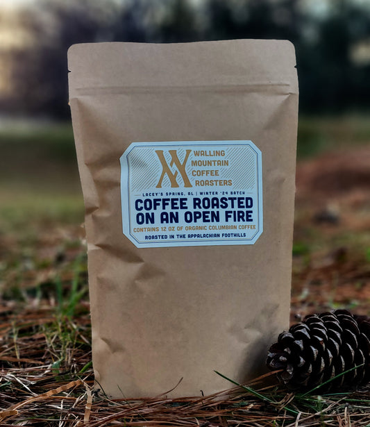 A brown bag of Walling Mtn. 12oz Whole Bean Coffee rests on pine needles with a pine cone, presenting an organic Colombian blend by Walling Mtn. Coffee, roasted using traditional methods.