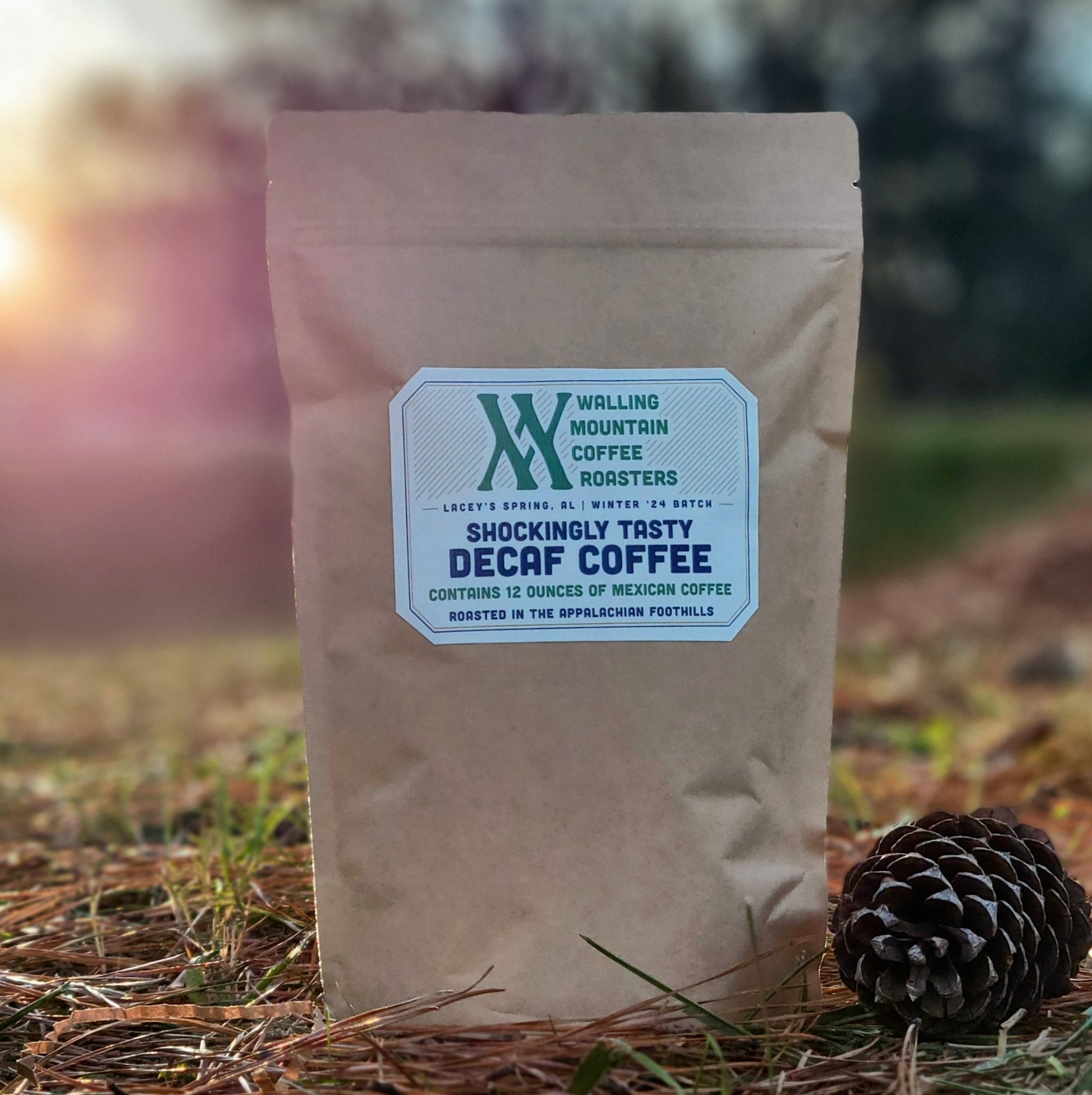 A 12oz bag of Walling Mtn. Coffee whole bean decaf sits on the ground near a pine cone, highlighting the rich aroma of their Organic Colombian/Peruvian Coffee Blend, crafted with traditional roasting methods against a beautifully blurred outdoor background.