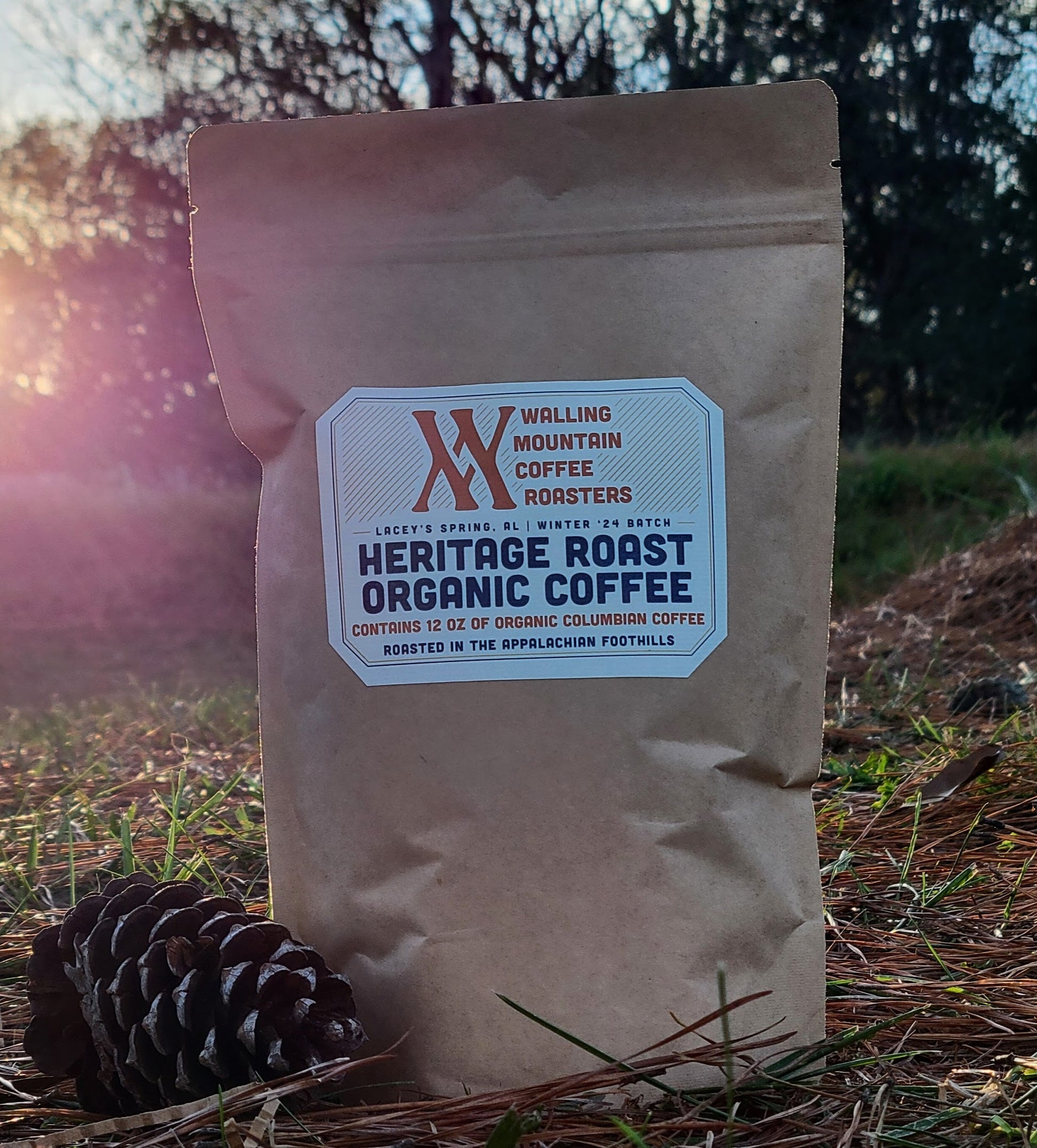 A 12oz bag of Walling Mtn. Coffee's whole bean Heritage Roast Organic Coffee rests by a pinecone against a sunset backdrop. This exquisite Colombian/Peruvian blend is crafted traditionally, offering a rich and authentic flavor experience.