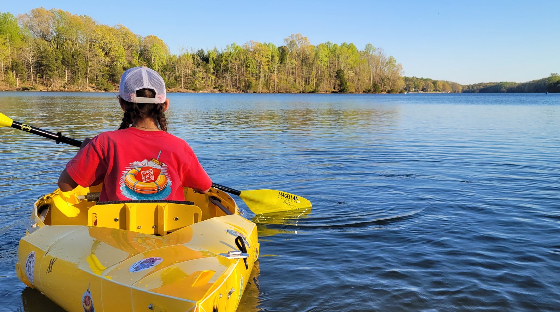 Wearing a Piper & Leaf Tea Co. Life Preserver T-Shirt and cap, a person paddles a yellow kayak on a calm lake, sipping iced tea, surrounded by trees and a clear sky.