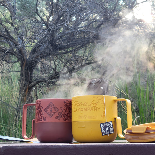 Two P&L 14oz Rise Mugs from Klean Kanteen by Piper & Leaf Tea Co.—one red, one yellow—made from recycled stainless steel sit outdoors on a table as steam rises from the yellow mug, with trees and grass behind.