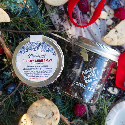 A Piper & Leaf Tea Co. Cherry Christmas Glass Jar of Loose Leaf Tea (30 servings) sits among pine branches, berries, cookies, and a red ribbon.