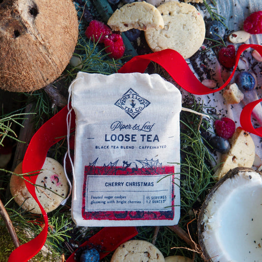 A Piper & Leaf Tea Co. Cherry Christmas Muslin Bag of Loose Leaf Tea (15 servings) sits among cookies, coconut halves, berries, evergreen branches, and a red ribbon—capturing the blend’s nutty hibiscus notes and festive warmth.