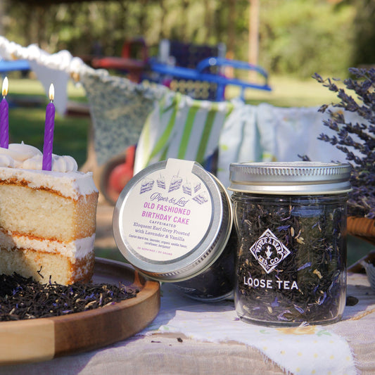 A slice of cake with two purple candles sits beside Piper & Leaf Tea Co.'s Old Fashioned Birthday Cake Glass Jar of Loose Leaf Tea (30 servings) on an outdoor table, with a blurred playground in the background.