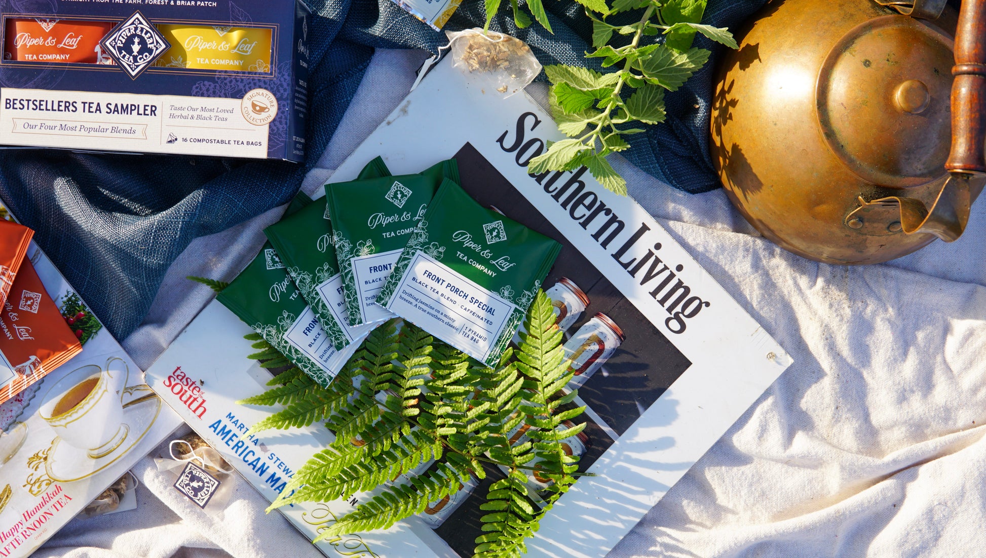 A Piper & Leaf Tea Co. Bestselling Tea Sampler (box of 16 tea bags), "Southern Living" magazine, green leaves, and a brass kettle rest on light fabric for a relaxed outdoor tea scene perfect for savoring an herbal infusion.